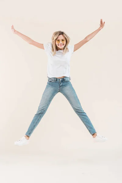 Happy young woman in eyeglasses jumping with raised arms isolated on beige — Stock Photo