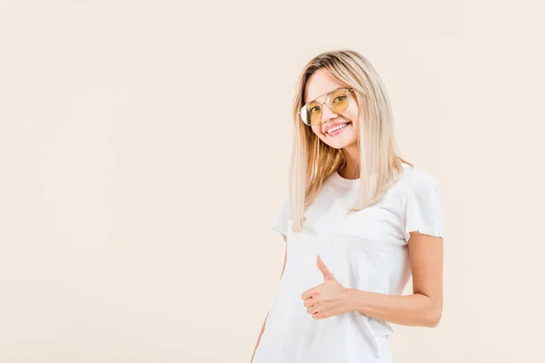 Happy young woman in sunglasses showing thumb up and smiling at camera isolated on beige — Stock Photo