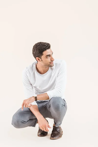 Handsome smiling young man crouching and looking away isolated on beige — Stock Photo