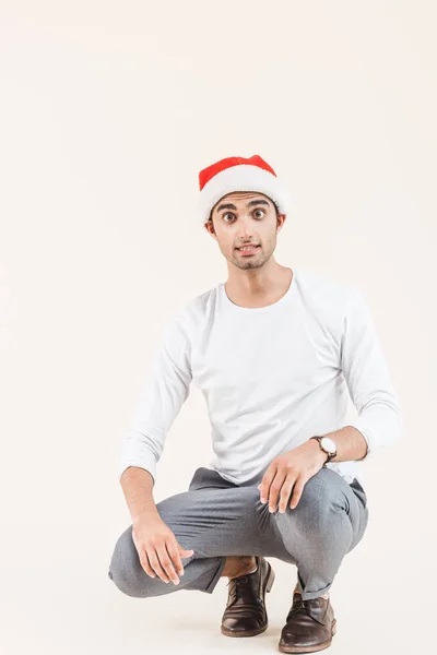 Scioccato giovane uomo in cappello di Babbo Natale accovacciato e guardando la fotocamera isolata sul beige — Foto stock