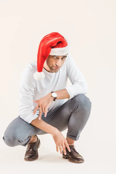 Giovane uomo in cappello di Babbo Natale accovacciato e guardando la fotocamera isolata sul beige — Foto stock