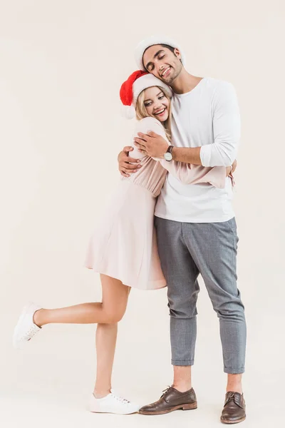 Feliz jovem casal em santa chapéus abraçando isolado no bege — Fotografia de Stock