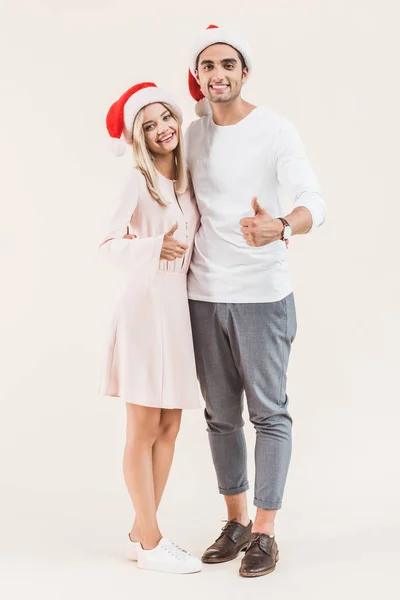 Feliz jovem casal em santa chapéus mostrando polegares para cima e sorrindo para a câmera isolada no bege — Fotografia de Stock