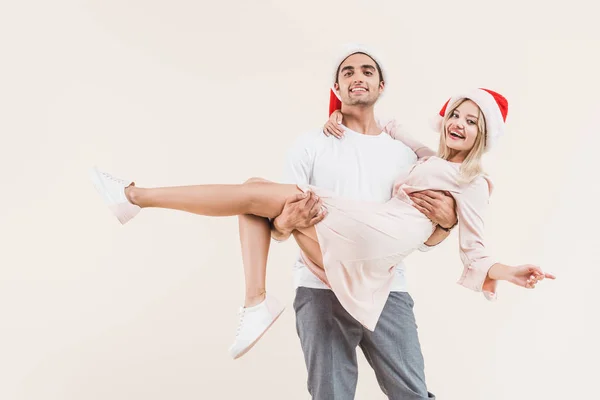 Glücklicher Mann mit Weihnachtsmannmütze, der eine schöne junge Frau trägt und isoliert auf Beige in die Kamera lächelt — Stockfoto