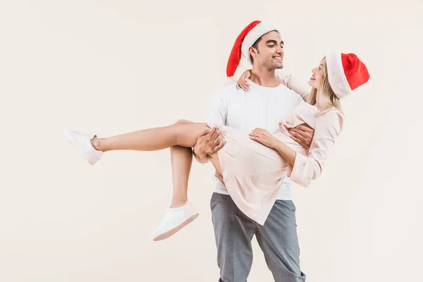 Happy young man in santa hat carrying beautiful smiling girl isolated on beige — Stock Photo