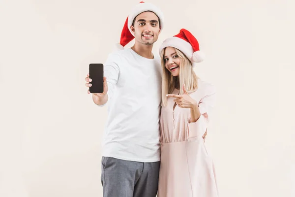 Feliz jovem casal em santa chapéus segurando smartphone com tela em branco e sorrindo para a câmera isolada no bege — Fotografia de Stock