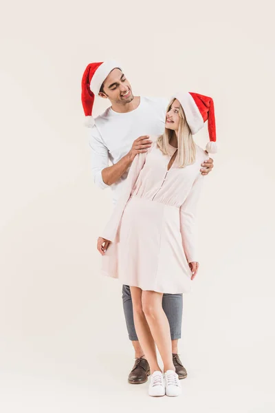 Ganzkörperansicht des schönen glücklichen jungen Paares in Weihnachtsmannmützen, die sich isoliert auf beige anlächeln — Stockfoto