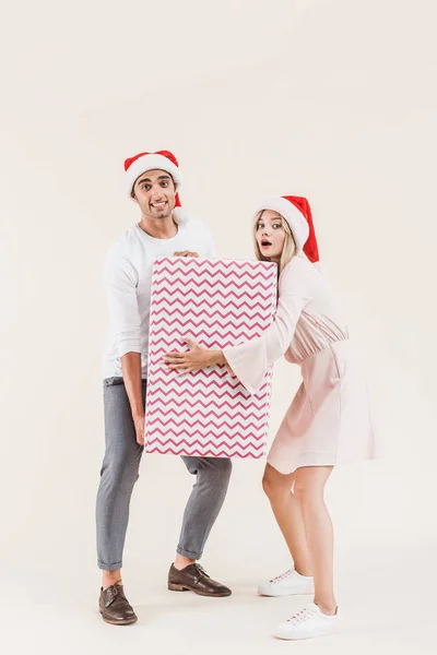 Junges Paar in Weihnachtsmannmützen mit großen Weihnachtsgeschenken und Blick in die Kamera isoliert auf beige — Stock Photo