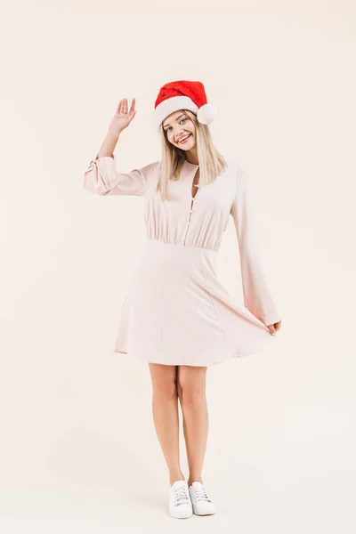 Piena vista lunghezza di bella felice giovane donna in abito e cappello di Babbo Natale sorridente alla fotocamera isolata sul beige — Foto stock