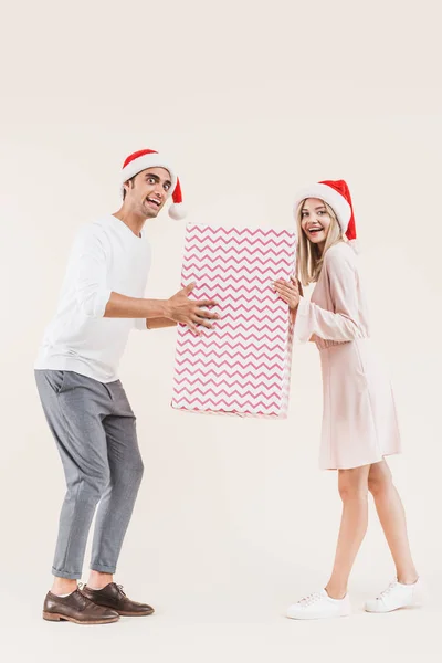 Feliz pareja joven en sombreros de santa celebración de regalo de Navidad grande y sonriendo a la cámara aislada en beige - foto de stock