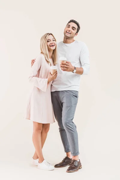 Feliz joven pareja sosteniendo vasos de papel y sonriendo a la cámara aislada en beige — Stock Photo