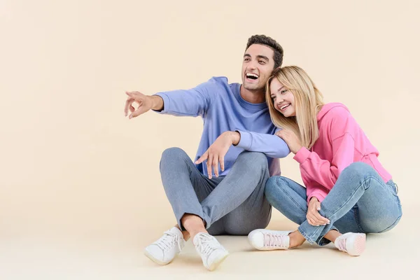 Joyeux jeune couple assis ensemble et pointant du doigt isolé sur beige — Photo de stock