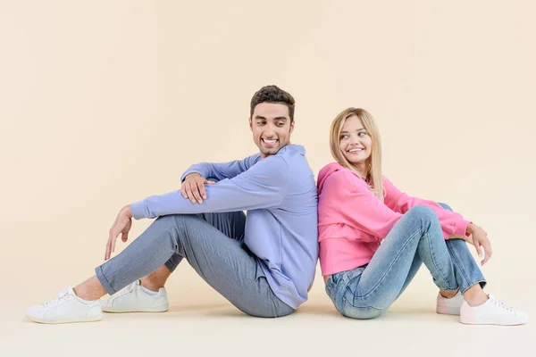 Vista lateral de feliz joven pareja sentada espalda con espalda aislada en beige - foto de stock