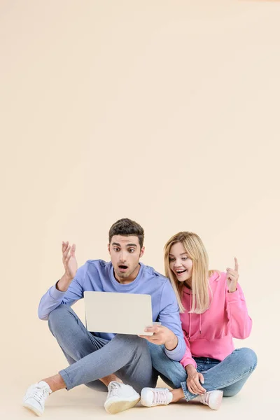 Überraschtes junges Paar sitzt zusammen und benutzt Laptop isoliert auf beige — Stockfoto