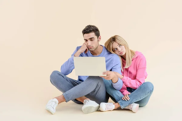 Gelangweiltes junges Paar sitzt zusammen und benutzt Laptop isoliert auf beige — Stockfoto