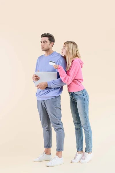 Young woman holding credit card and looking at boyfriend with laptop isolated on beige — Stock Photo