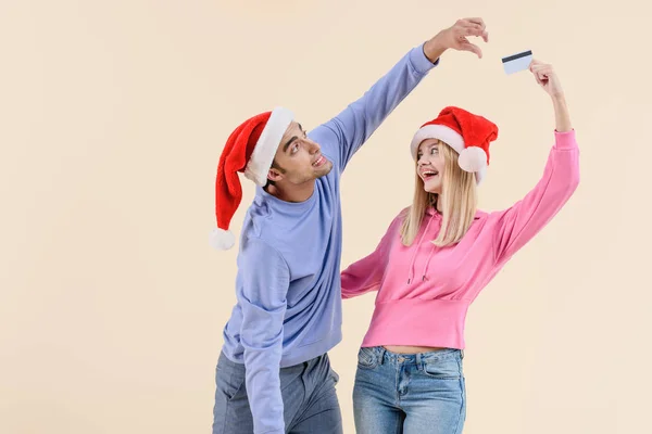 Giovane coppia in cappelli di Babbo Natale divertirsi con carta di credito isolata sul beige — Foto stock