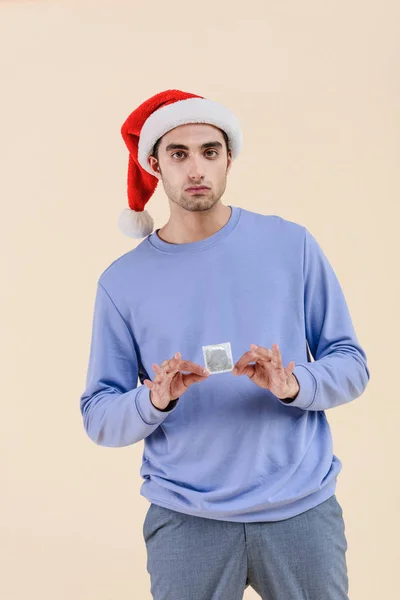 Красивий молодий чоловік у капелюсі Санта тримає презерватив і дивиться на камеру ізольовано на бежевому — стокове фото