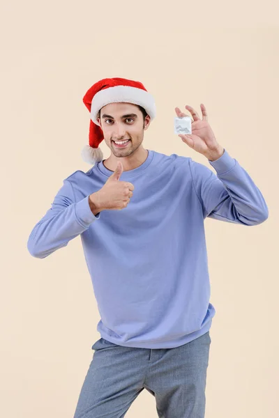 Glücklicher junger Mann mit Weihnachtsmütze, Kondom in der Hand und Daumen hoch auf beige — Stockfoto