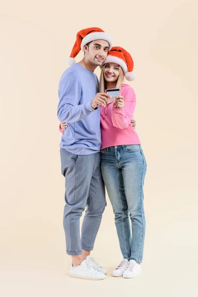 Feliz pareja joven en sombreros de santa celebración de la tarjeta de crédito y sonriendo a la cámara aislada en beige - foto de stock