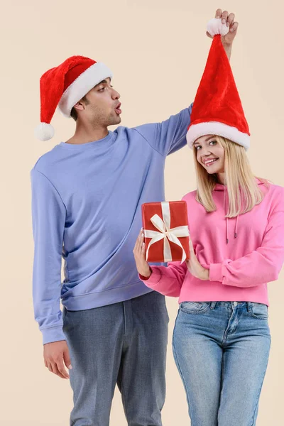 Feliz joven mujer sosteniendo regalo de Navidad y sonriendo a la cámara mientras el hombre se divierte con sombrero de santa aislado en beige - foto de stock