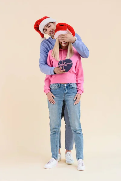 Sonriente joven hombre cerrando los ojos a la mujer feliz y presentando caja de regalo en forma de corazón aislado en beige - foto de stock