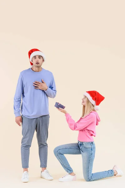 Lächelnde junge Frau kniet und überreicht Geschenkbox an schockierten jungen Mann mit Weihnachtsmann isoliert auf beige — Stockfoto