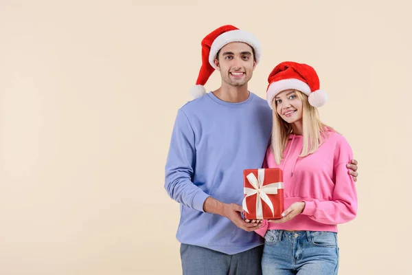 Glückliches Paar in Weihnachtsmützen mit Weihnachtsgeschenk und lächelnd in die Kamera isoliert auf beige — Stockfoto