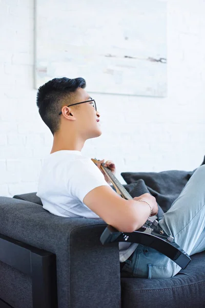 Side view of asian guitarist playing electric guitar on sofa at home — Stock Photo