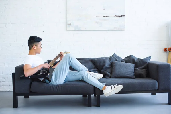Seitenansicht eines hübschen asiatischen Mannes, der auf dem Sofa liegt und zu Hause unplugged E-Gitarre spielt — Stockfoto