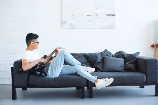 Vue latérale du bel homme asiatique couché sur le canapé et jouant de la guitare électrique débranchée à la maison — Photo de stock