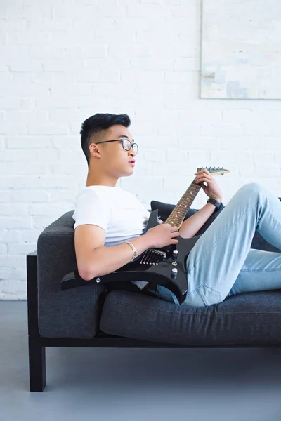 Beau asiatique homme couché sur canapé et jouer unplugged guitare électrique à la maison — Photo de stock