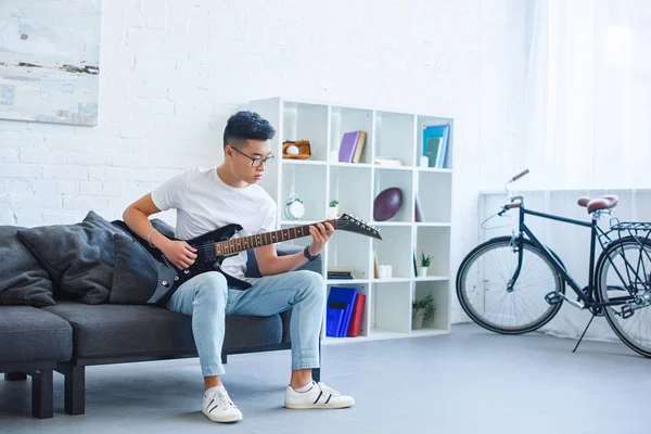 Bello asiatico uomo giocare nero elettrico chitarra su divano a casa — Foto stock