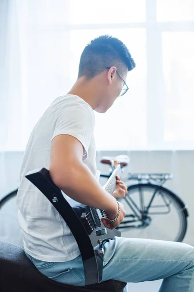 Vista lateral del hombre asiático tocando la guitarra eléctrica desenchufada en casa - foto de stock