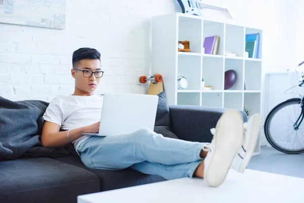 Шокований красивий азіатський чоловік використовує ноутбук на дивані вдома — стокове фото