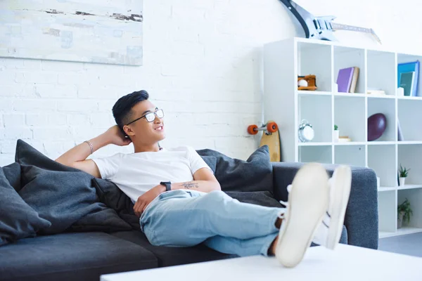 Smiling handsome asian man sitting on sofa with legs on table at home, looking away — Stock Photo