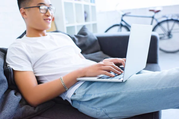 Vista laterale di allegro bello asiatico uomo utilizzando laptop su divano a casa — Foto stock