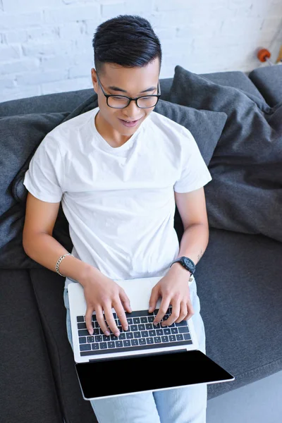 Alto angolo vista di bello asiatico uomo utilizzando laptop su divano a casa — Foto stock