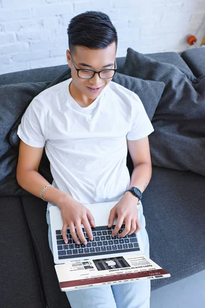 Hochwinkel-Ansicht eines hübschen asiatischen Mannes mit Laptop mit geladenen bbc Nachrichtenseite auf dem Sofa zu Hause — Stockfoto