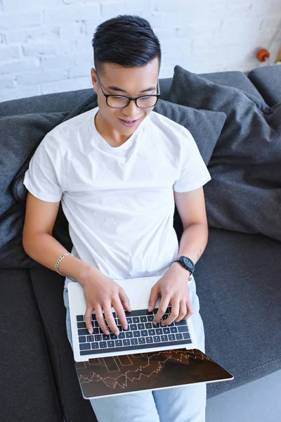 Vista ad alto angolo di bell'uomo asiatico utilizzando laptop con elettrodomestico grafico sul divano a casa — Foto stock