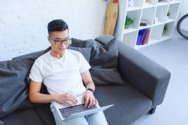 Alto ângulo vista de bonito alegre asiático homem usando laptop no sofá em casa — Fotografia de Stock