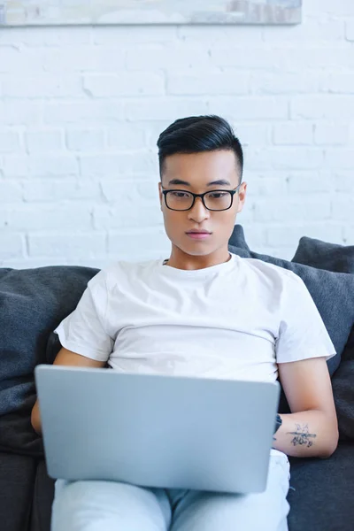 Bel homme asiatique dans des lunettes en utilisant un ordinateur portable sur le canapé à la maison — Photo de stock