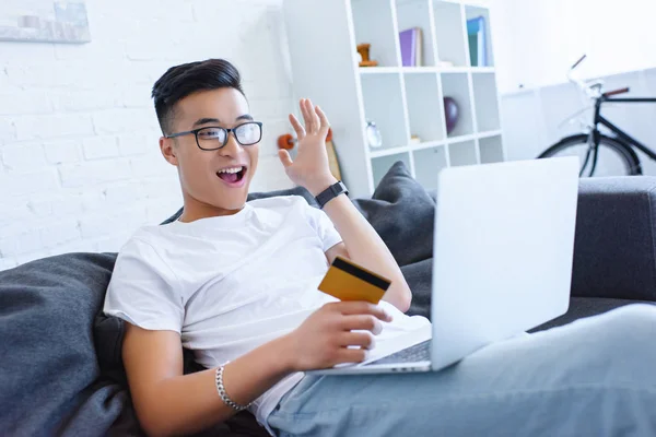 Happy handsome asian man using laptop and holding credit card on sofa at home — Stock Photo