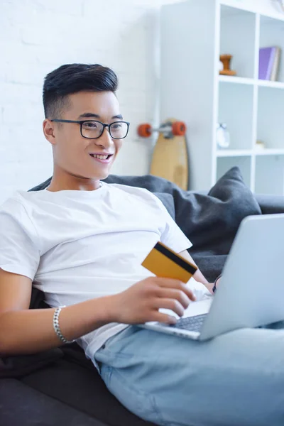 Sorrindo bonito asiático homem usando laptop e segurando cartão de crédito no sofá em casa — Fotografia de Stock