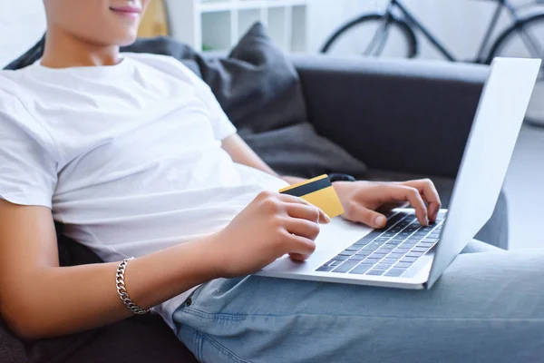 Imagem cortada do homem em roupas casuais usando laptop e segurando cartão de crédito no sofá em casa — Fotografia de Stock
