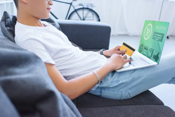 Immagine ritagliata dell'uomo che usa il computer portatile con l'apparecchio di shopping e che tiene la carta di credito sul sofà a casa — Foto stock