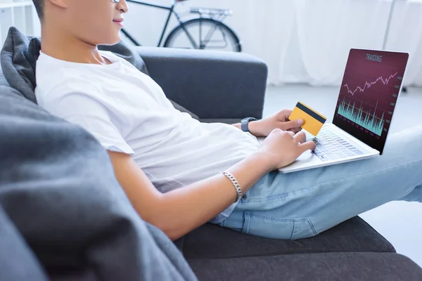 Cropped image of man using laptop with traiding appliance and holding credit card on sofa at home — Stock Photo