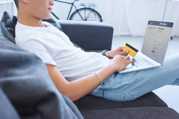 Immagine ritagliata dell'uomo con computer portatile con pagina facebook caricata sul divano e con carta di credito in mano a casa — Foto stock