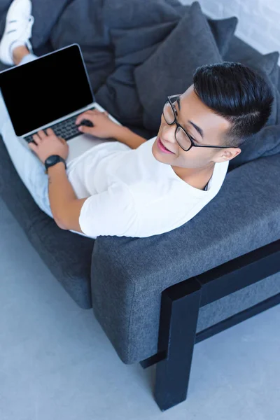 Ângulo alto vista de bonito asiático homem deitado no sofá e usando laptop com tela em branco em casa — Fotografia de Stock