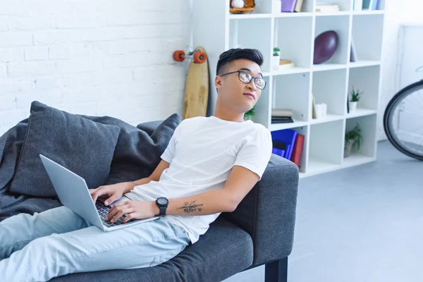 Bello asiatico uomo utilizzando laptop su divano a casa e guardando lontano — Foto stock